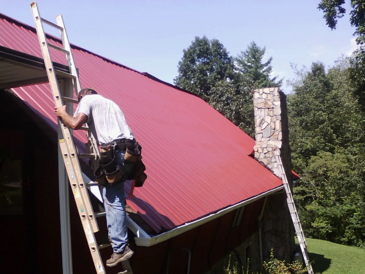 Roofer on a ladder beside a red metal roof and stone chimney — Soffit & Fascia Repair by Roofing Brothers in Fall Creek